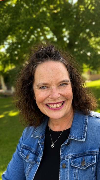 Jenny Mills smiling in a denim jacket under a shade tree